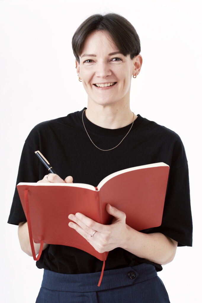 An image of poet Alice Frecknall. She has short black hair, she wears a black t-shirt and blue trousers. She smiles at the camera and holds a black pen and red writing book.