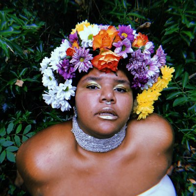 A woman with large floral wreath around her head looks into the camera wearing a choker necklace and blue lipstick
