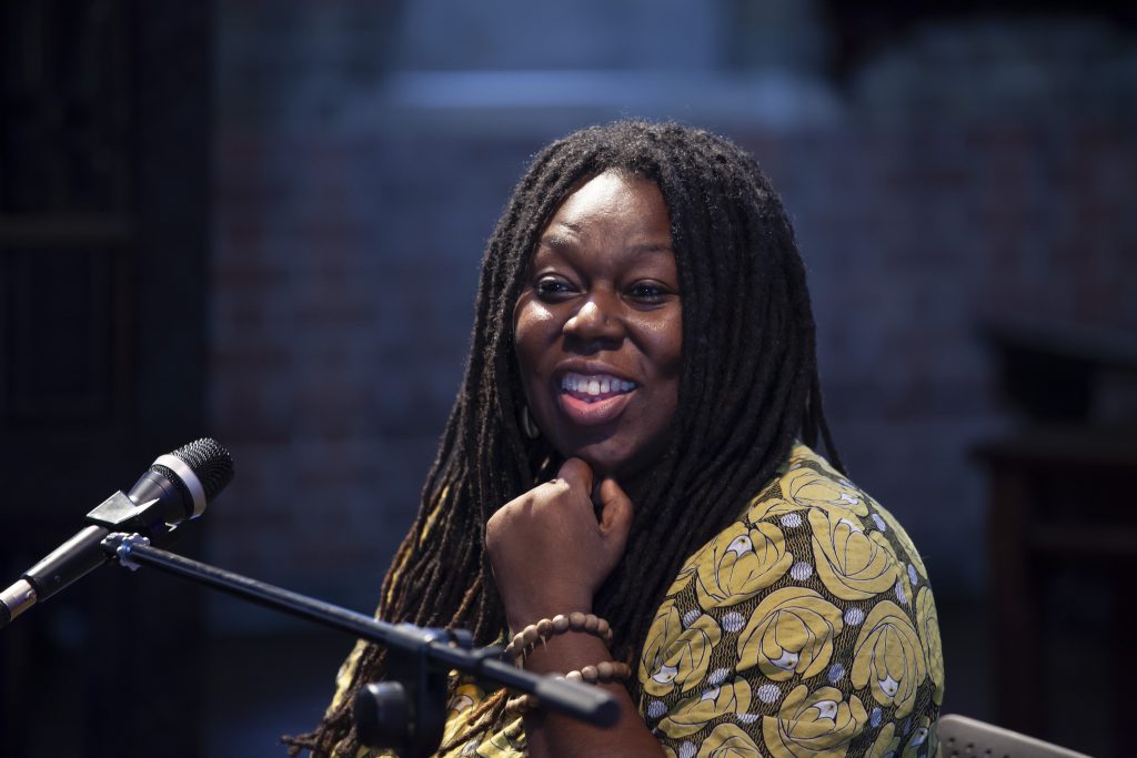 Woman with patterned top and dreadlocks speaking into a microphone