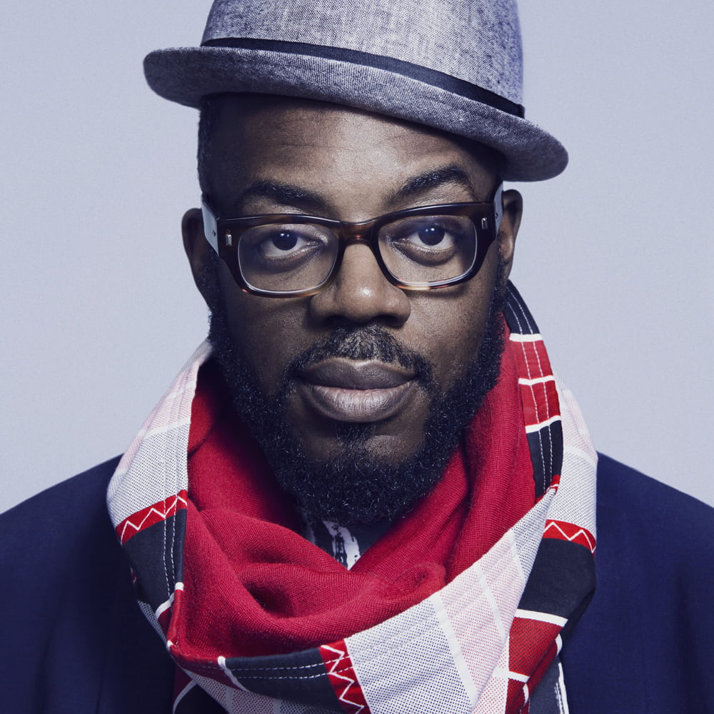 Breis wears a grey bowler hat, red white and blue scarf, with thick rimmed glasses.