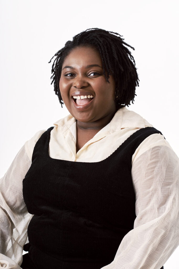 An image of poet Eileen Gbagbo. She smiles at the camera and has short black hair. She wears a black dress and white shirt.