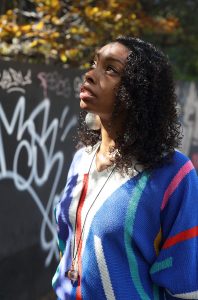 A woman with mid lenght, curly hair looks up into the shaded trees. She is wearing a verticle striped jumper in primary colour shades