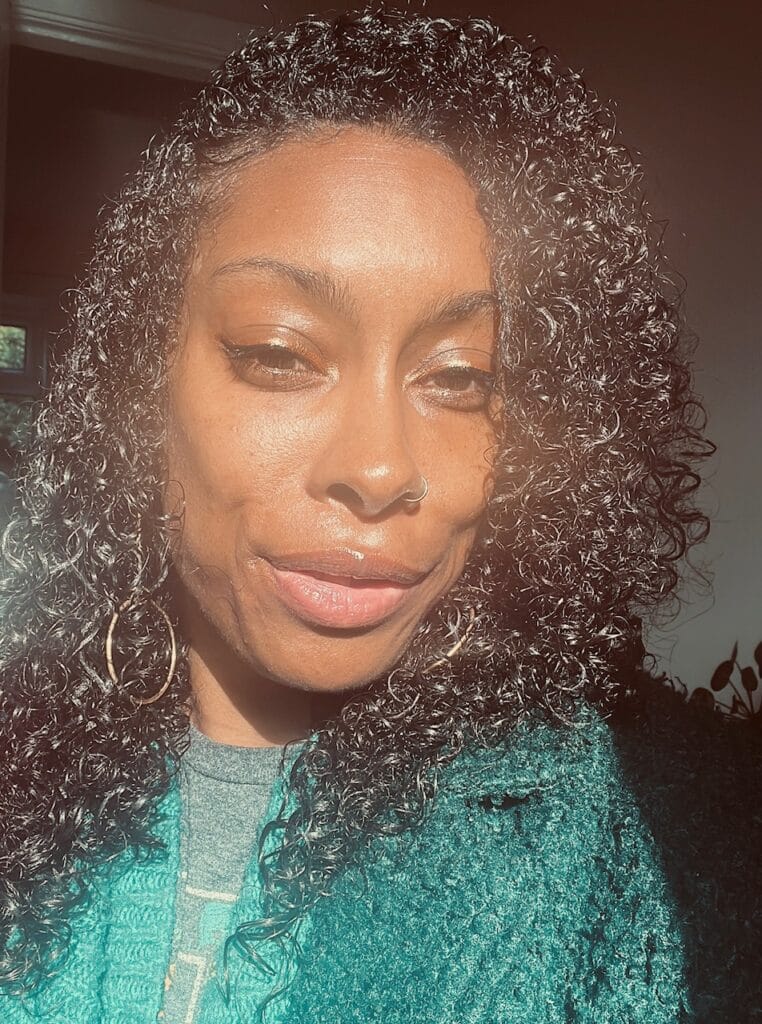 An image of poet Ioney Smallhorne, she has long, black hair, down to her shoulders. Wears gold hooped earrings and a green knitted cardigan, underneath this is a grey t-shirt. 