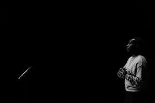 A photo of poet Jerome Scott performing. he wears a white t-shirt and is positioned in the left hand corner of the image. The rest of the image is filled with the dark black of the unlit stage.