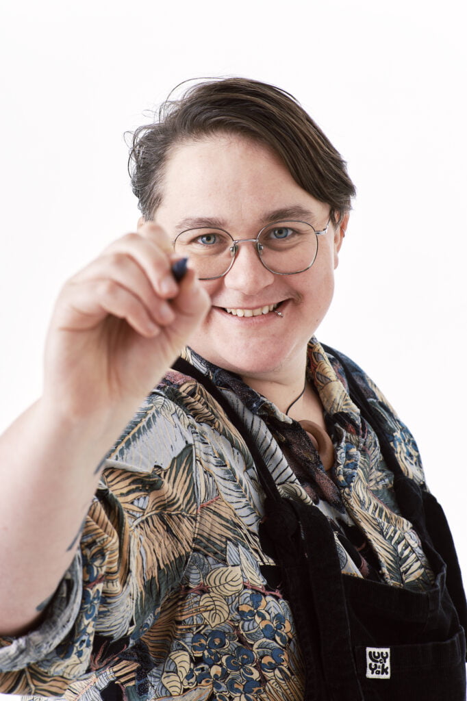 an image of poet Linden McMahon. They has short brown hair and glasses. They wear black dungarees and a brown and grey shirt. They hold up a pen towards the camera.