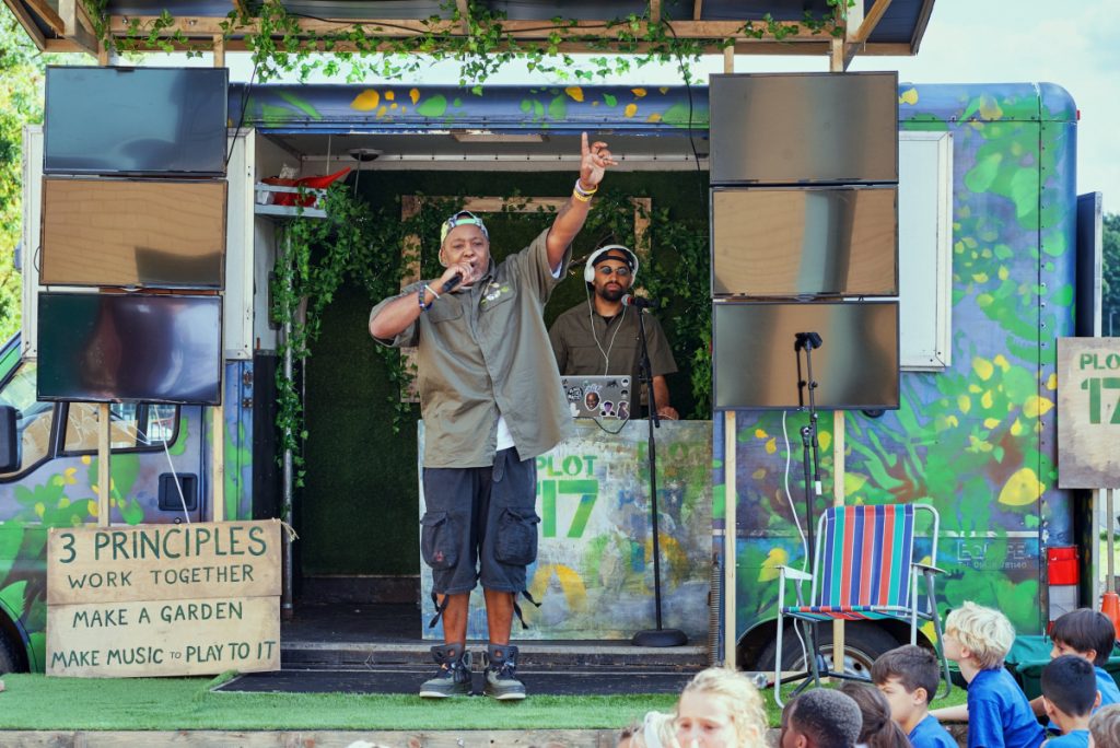 Image of a man with shorts a shirt and a backwards cap standing on stage with his arm raised