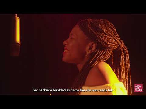Screenshot from a video of Safiya speaking into a microphone in a dark room with orange and yellow lighting