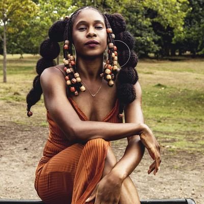 A woman with hair tied in bubbled bunches, heavy with beads wearing an orange jumpsuit kneels on the ground outisde