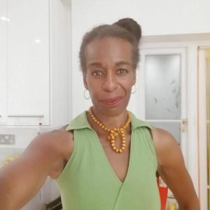 A woman in a green sleeveless top and yellow beaded necklace with her hair in a bun smiles at the camera