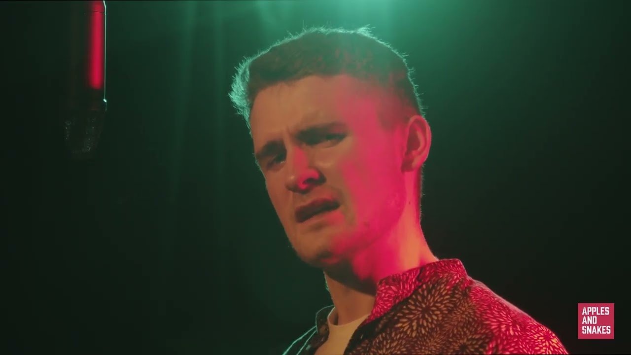 Screenshot from a video of Shaun Hill speaking in to a microphone in a dark room with green and red lighting