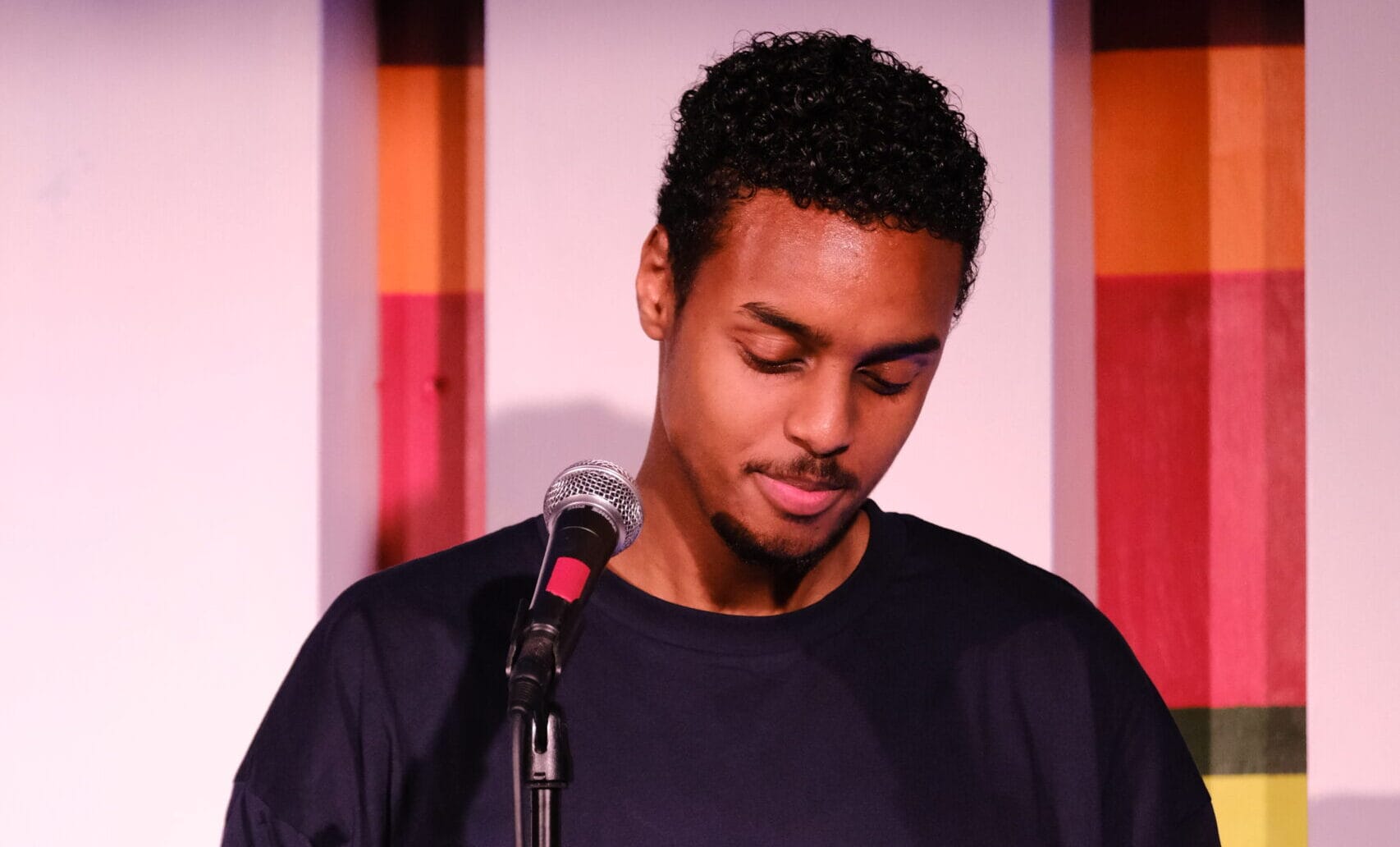 Poet Zakariye is standing at a microphone reading a book. He wears a black t-shirt and has short black hair.