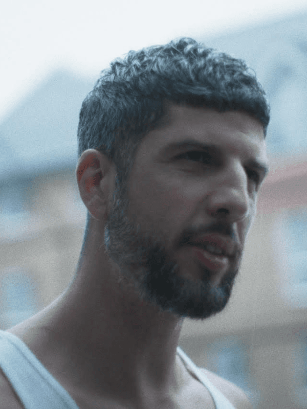 Antony looks on intensely wearing a sleeveless white vest, cropped hair and short beard