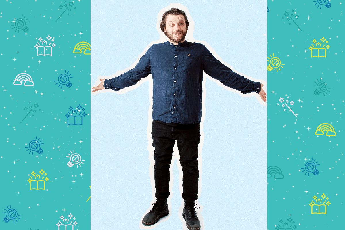 An image of poet Adam Kammerling. He wears a blue shirt and black trousers, he wears black trainers. His arms are outstretched to each side of his body as he smiles at the camera. He has long shoulder length brown hair and a brown beard.