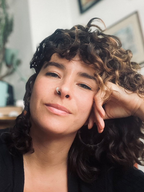 Amy leans head against hand, curly brown hair flowing over hand, ring nose piercing with dark top