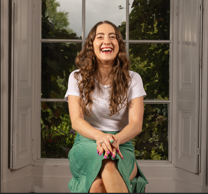 An image of poet Beth Kelly. She sits in front of a window and crosses her legs, her hands are placed on her knees.  She wears a green skirt and white t-shirt. She smiles towards the camera. She has long, brown, curly hair and pink fingernails. She has a green ring on her Index finer.