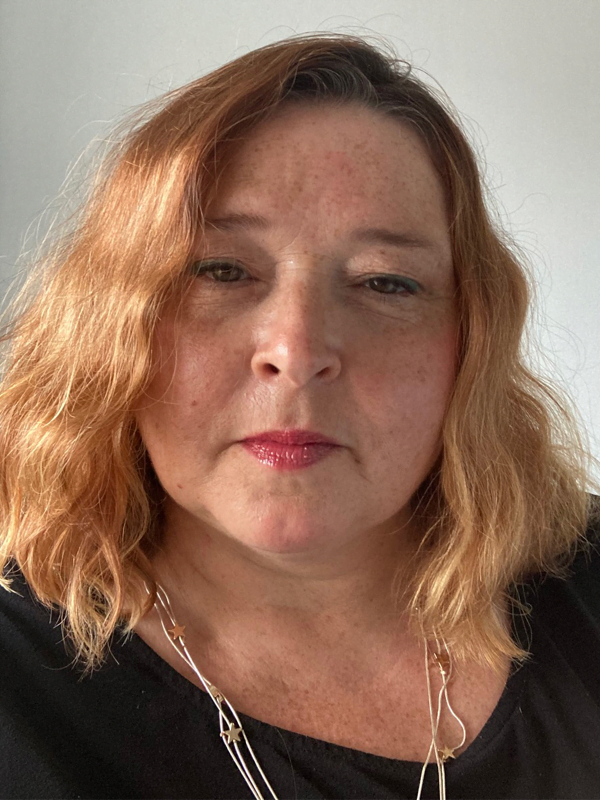 Charlotte wears a silver starred necklace, with black top. Hair is shoulder length, wavy and strawberry blonde.