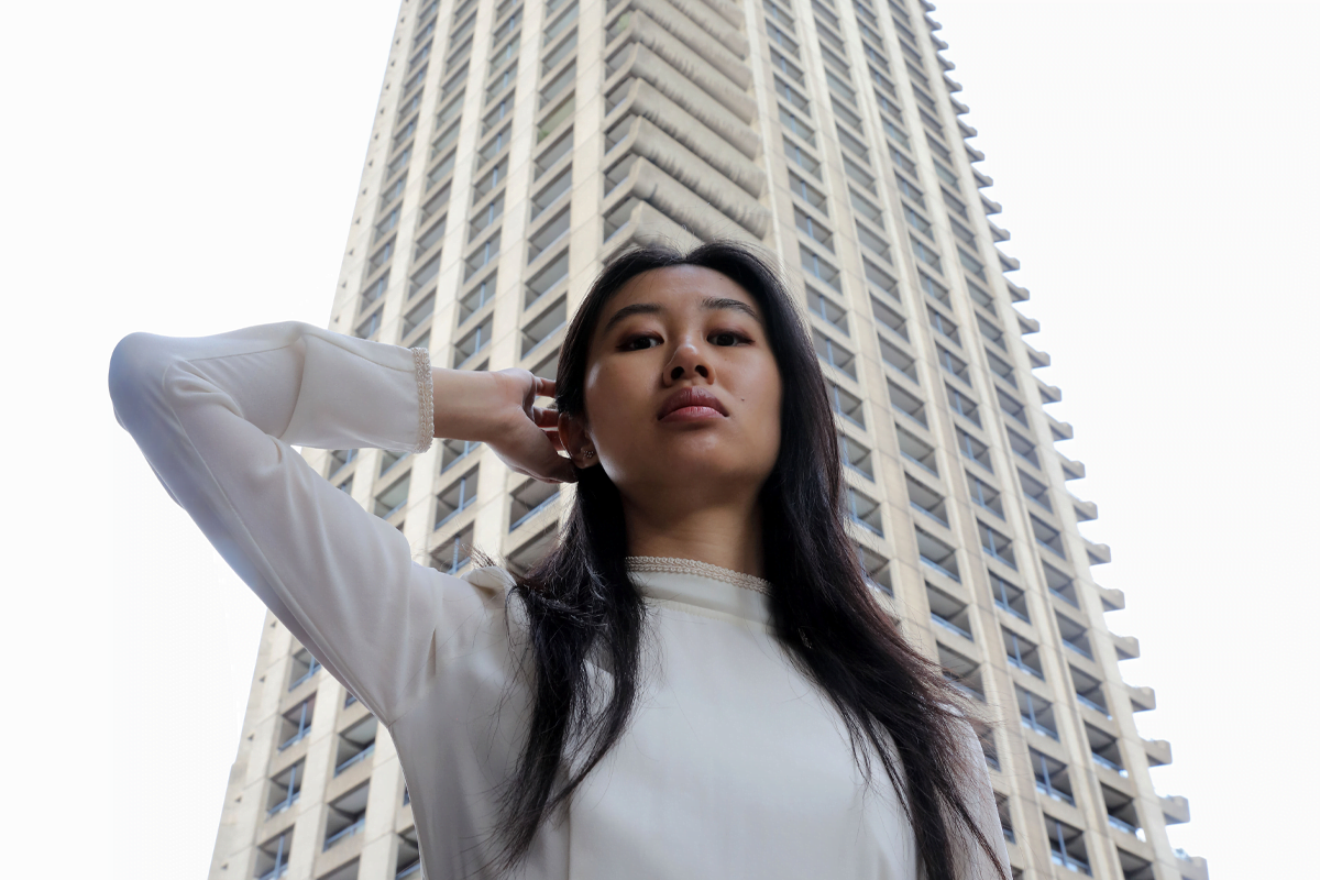 A woman with long dark hair wearing a high necked, long sleeve white dress puts her arm behind her head, standing in front of a tall tower block