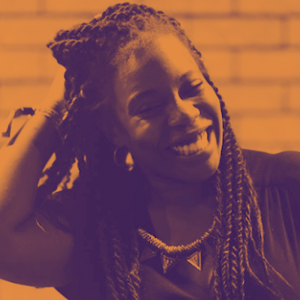 A woman with long braided hair and ornate necklace, runs her hand through the back of her hair and smiles happily