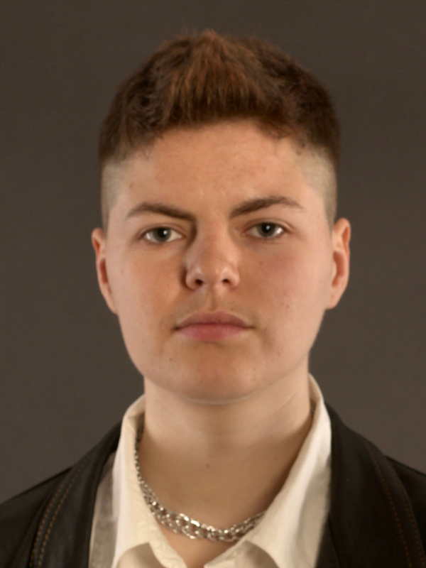 Eyrn has short, spiked up hair. Wearing a silver chain necklace, white shirt and leather jacket