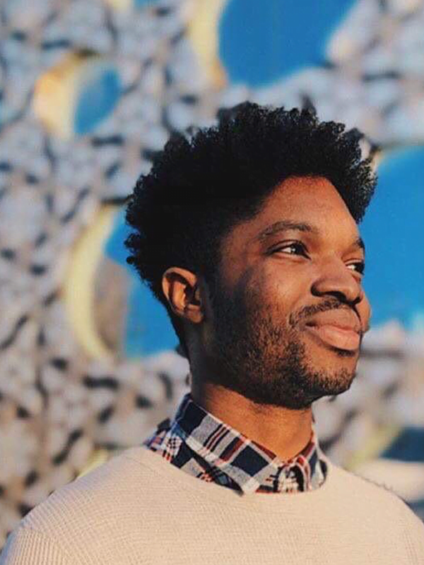 Isaac looks sideways, with short afro hair and shaved beard. Wearing a tartan shirt under a cream jumper, against floral and blue backgroud