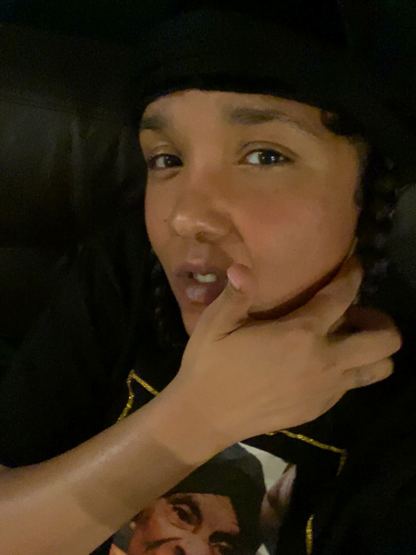 Jaidah holds hand over face, wearing a portrait shirt and black hat