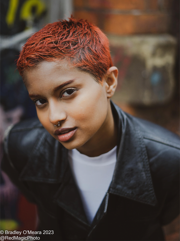 Jay has short, bright orangey red hair with nose piercing. Wearing a leather jacked and white t-shirt