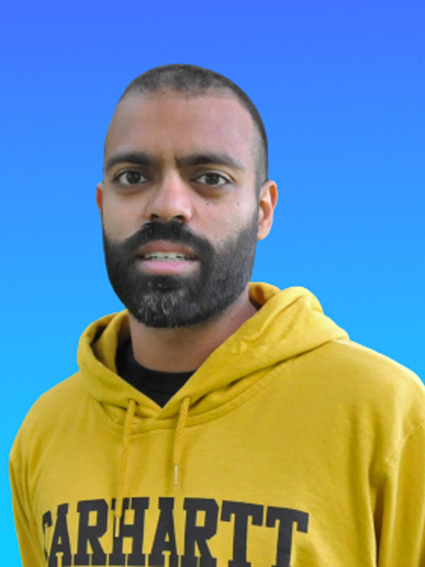 Jay is wearing a yellow carhartt hoodie, with shaved head and short shaved beard on a blue background