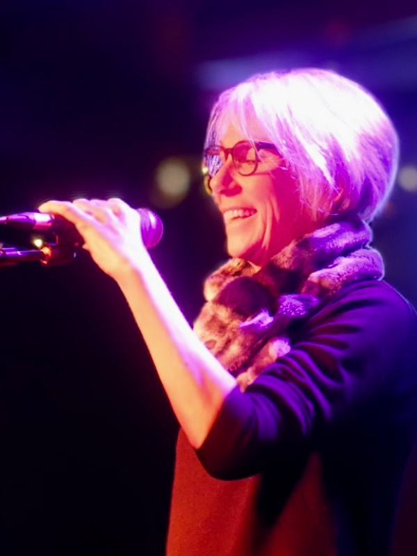 Jo has bobbed silver hair, wearing a animal print scarf, black cardigan and red top - about to talk into the mic