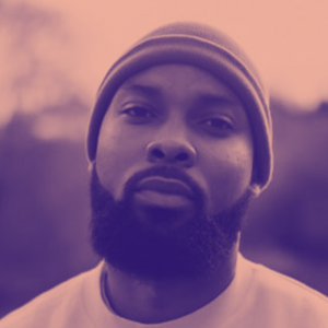 A man with a shapely beard and small knitted hat looks on with a neutral expression