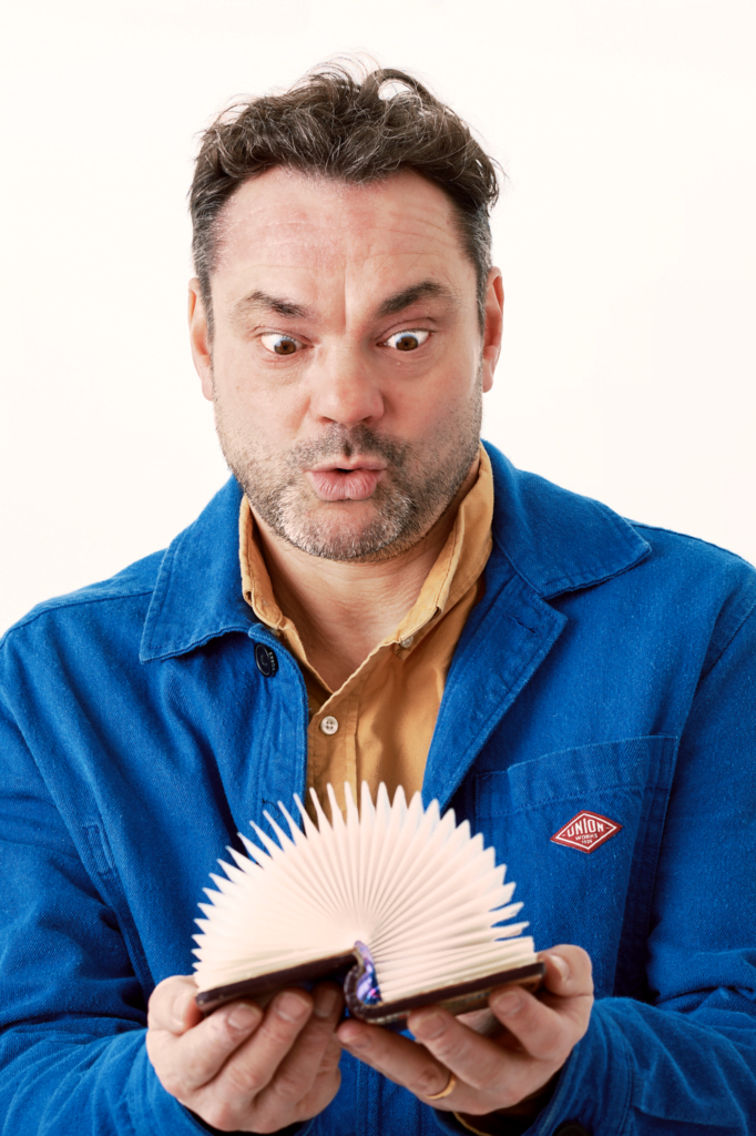 An image of poet Justin Coe. Justin has short brown hair zinc wears a blue jacket and brown shirt. He looks at a pop up book he is holding.