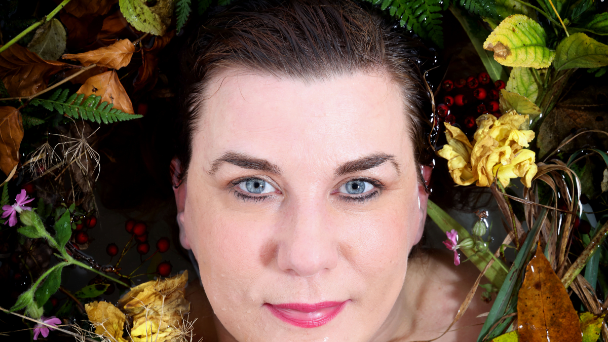 A woman with short dark hair and blue eyes surrounded by leaves and flowers