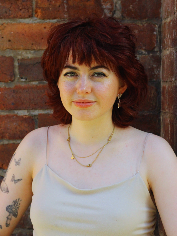 Lauren has a dark red mullet, wearing a white vest top with gold star necklace and earrings. Tattoos of butterflies and mushrooms on the left arm.