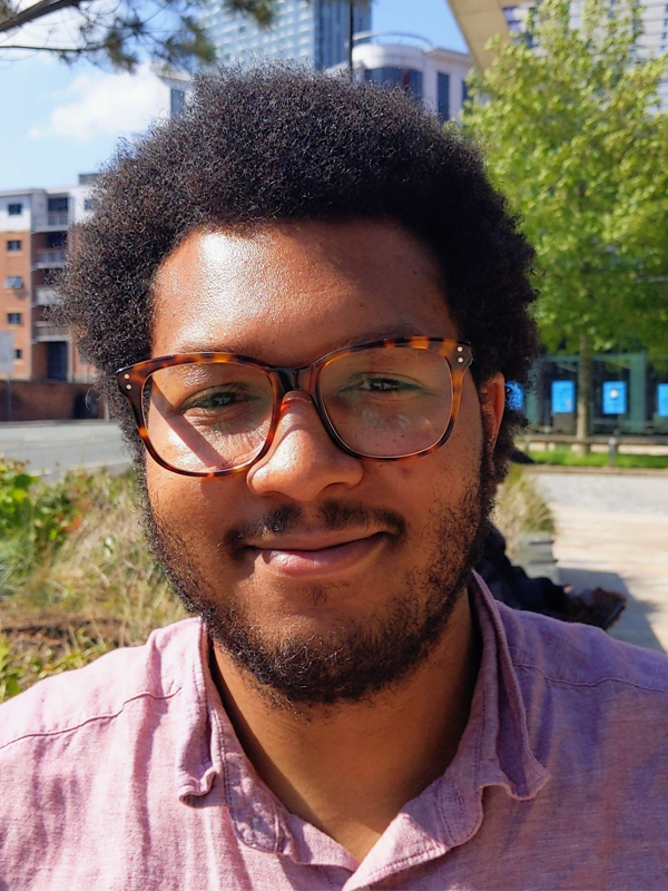 Louis has a short afro and dark short beard, wearing tortoiseshell glasses and a purple lined shirt