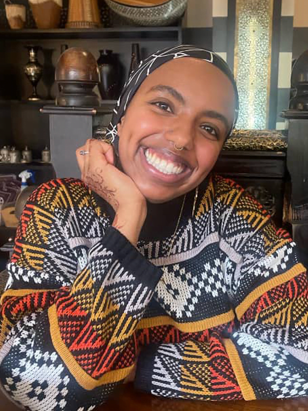 Milga smiling, wearing a black and white headband. and black, white, orange and red ethnic patterned jumper