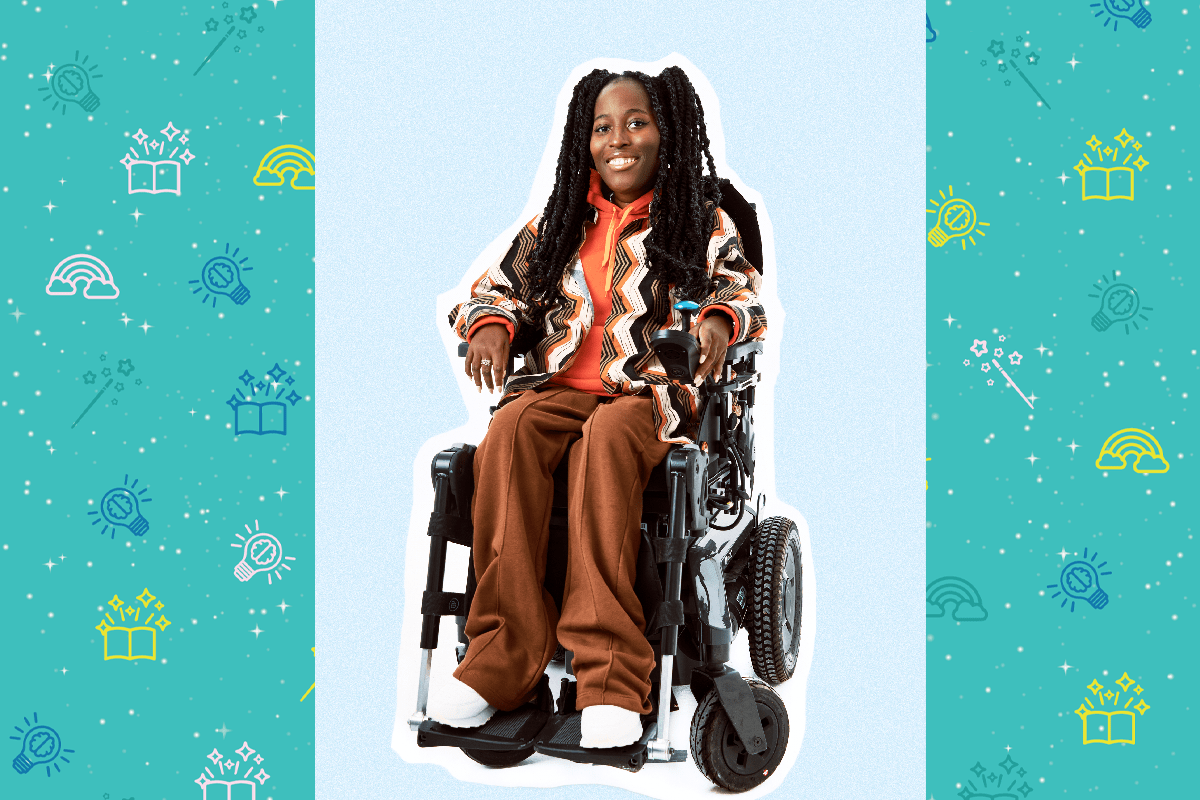 An image of Poet Miss Jacqui, She has long black hair and wears an orange hoodie, she also has a brown, orange and white coat on, she wears brown trousers. She smiles at the camera, her left hand rests on the blue controls of her wheelchair.