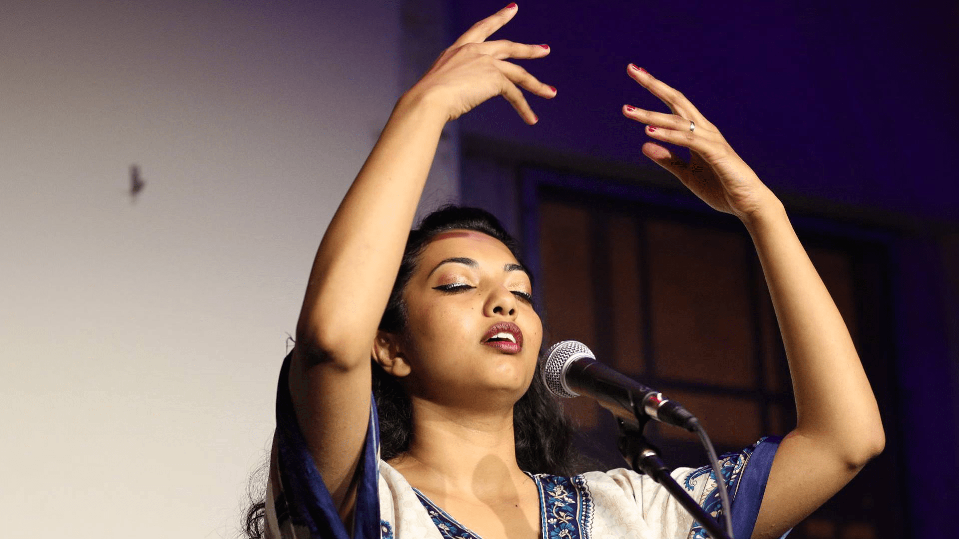 A woman with tied back dark hair and painted lips, raises her arms whilst speaking into a mic on stafe