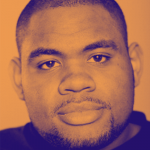 A man with a short trimmed beard and shaved head looks onto the camera