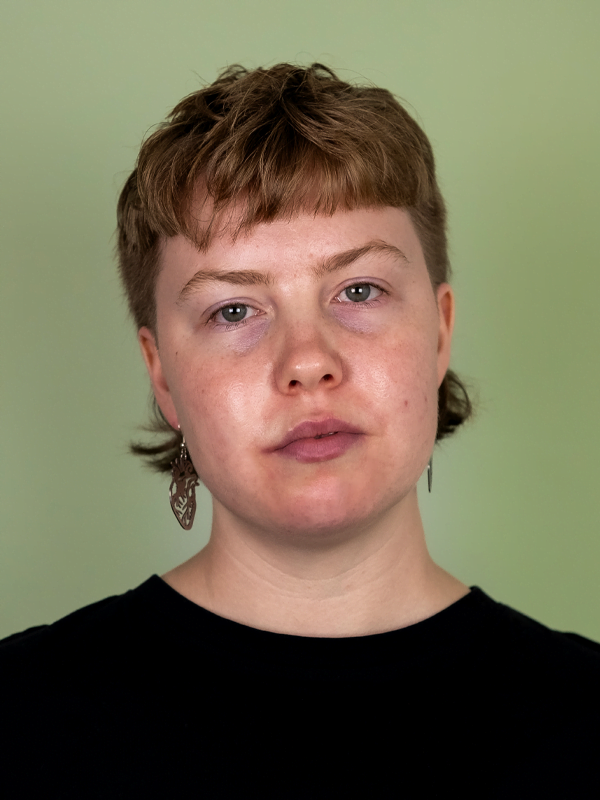 Sofe has a short mullet, wearing anatomical heart earrings, black t-shirt on a green background