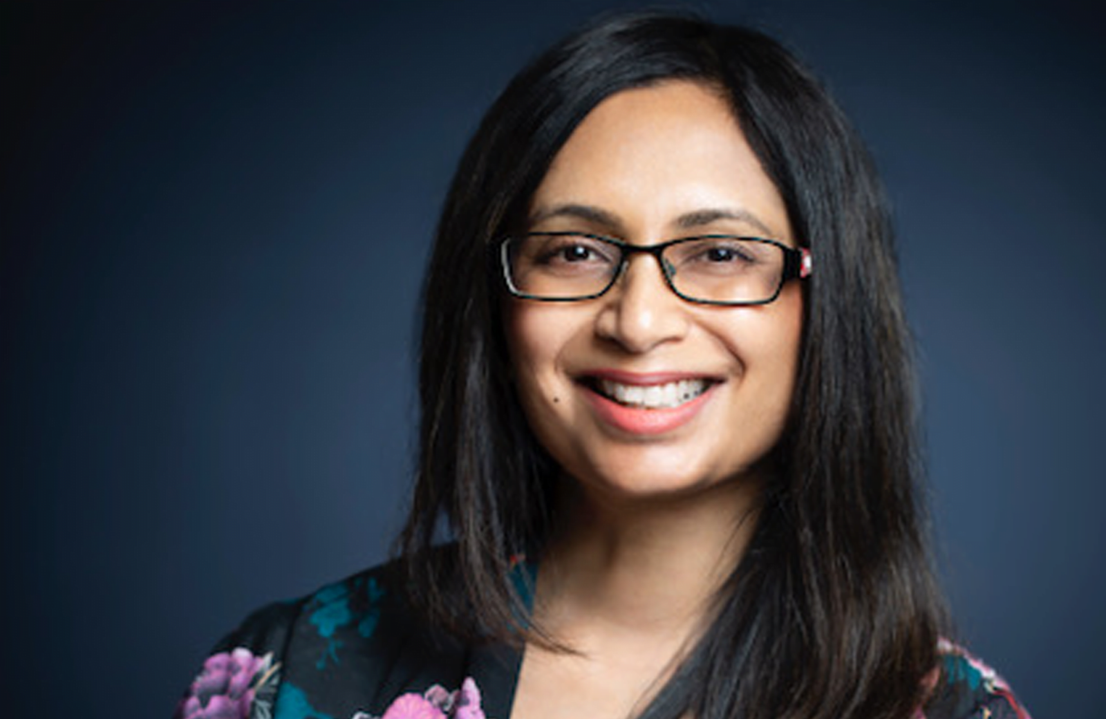 A woman with long dark hair, centre parted, wearing black rimmed glasses, smiling.
